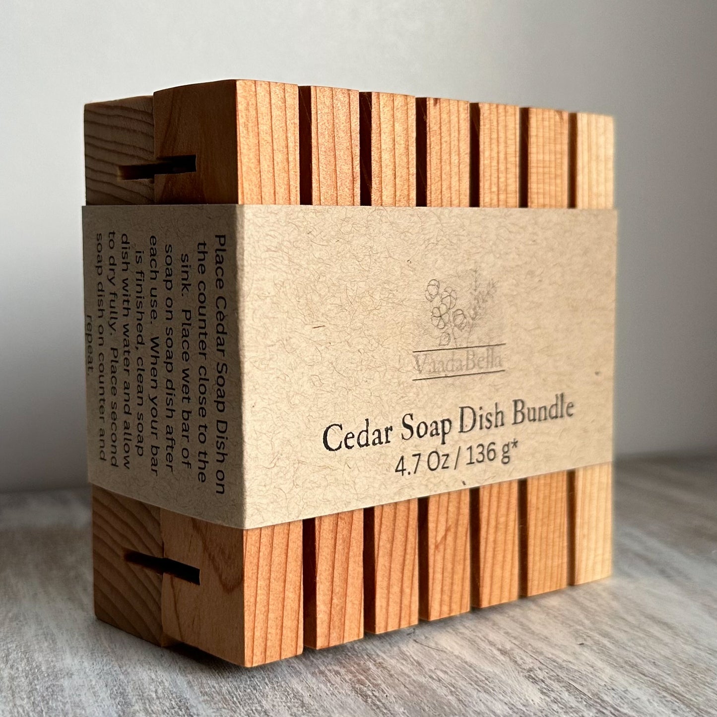 Two cedar planks with slots bundled together with brown kraft paper sleeve centered around the planks. Black ink print reads "Cedar Soap Dish Bundle, 4.7 oz / 136 g*"