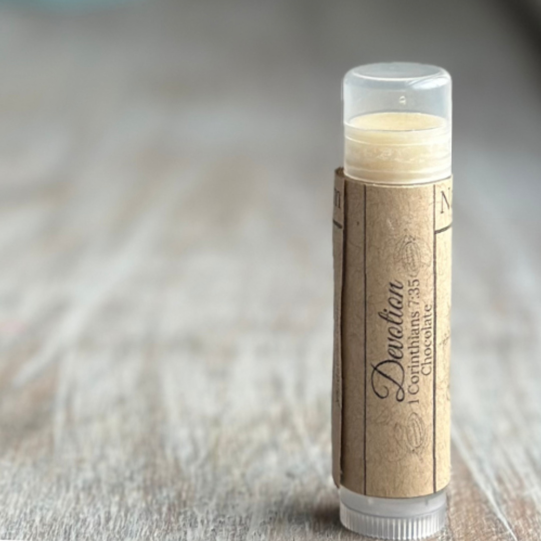 Stick of buttery yellow lip balm standing verticle on a weathered wood background, all on the right side of the image. Kraft paper label with "Devotion", 1 Corinthians 7:35 and "chocolate" printed in black.