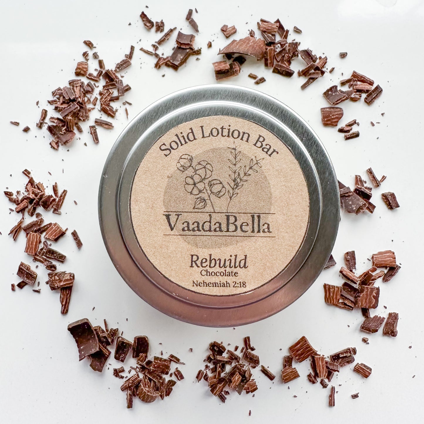 Front view of a circular silver metallic tin with lid. brown kraft paper circle label centered. on the top, Logo, "Solid Lotion Bar" "Rebuild" "Nehemiah 2:18" "Chocolate" is all printed in black ink. Chocolate shavings scattered around the tin.