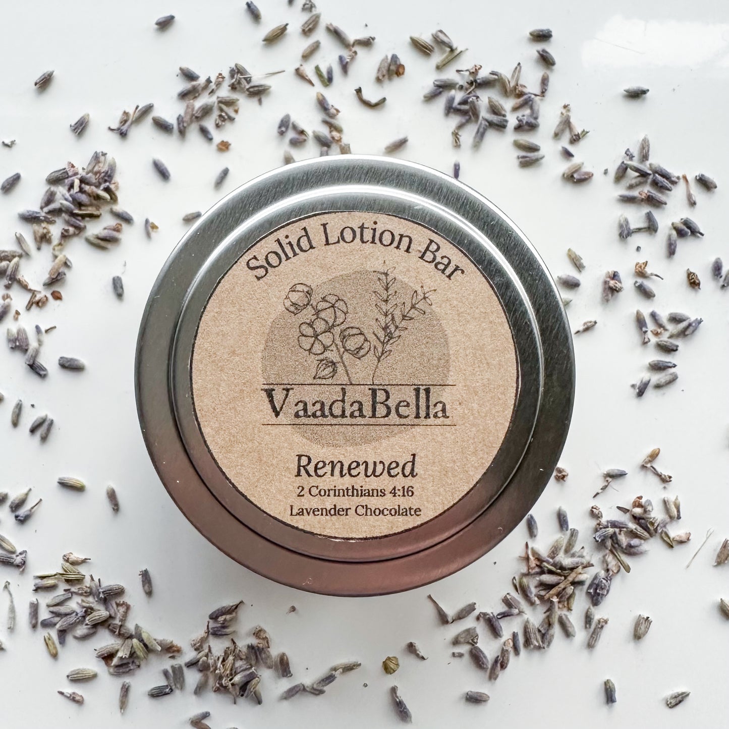 Bird's-eye view of a circular silver metallic tin with lid. brown kraft paper circle label centered. on the top, Logo, "Solid Lotion Bar" "Renewed" "2 Corinthians 4:16" "Lavender Chocolate" is all printed in black ink. Lavender buds sprinkled around the tin.