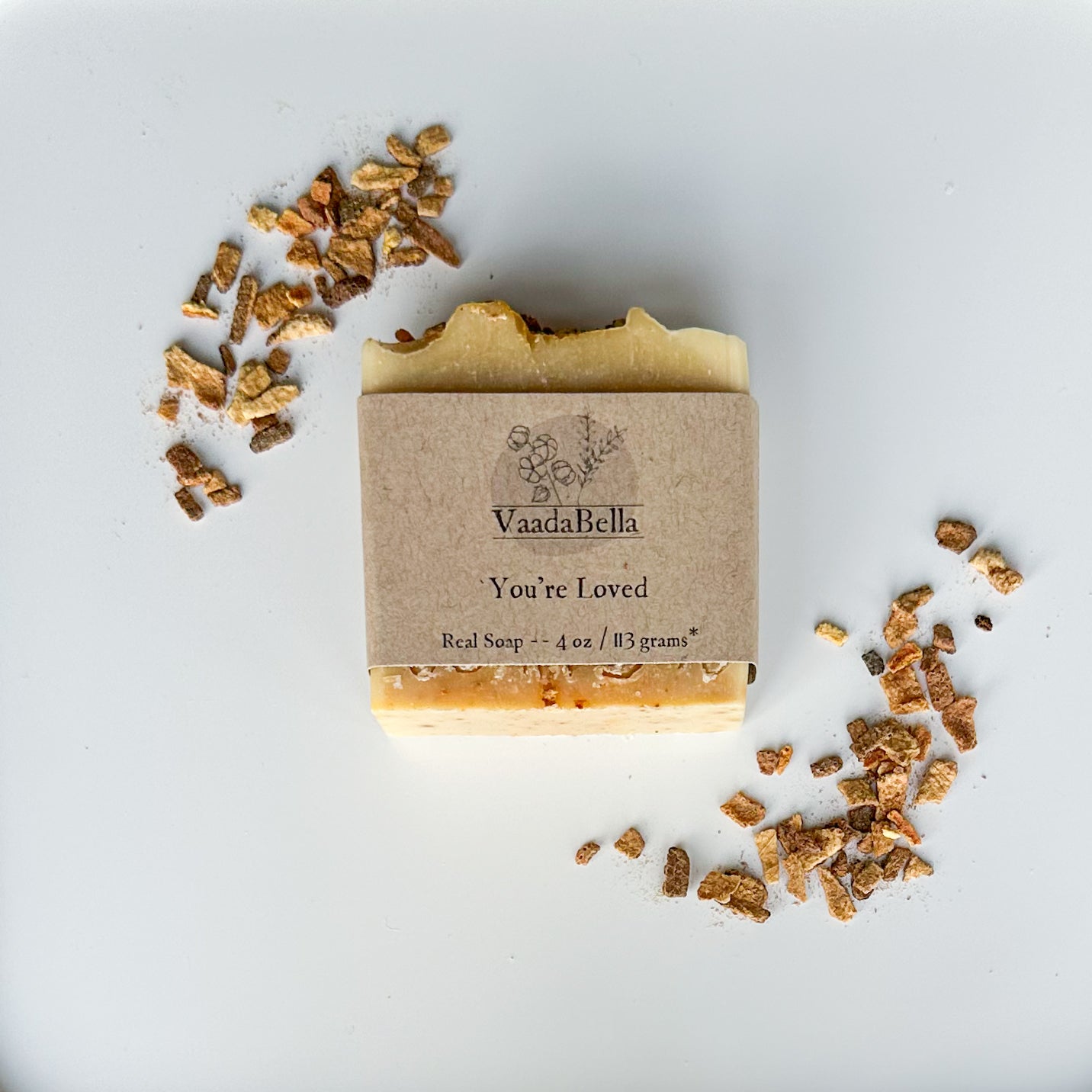 Yellow orange bar with slightly lighter top with kraft paper product sleeve centered on bar. Black ink prints the brand logo, "You're Loved" and "Real Soap -- 4 oz / 113 grams*"