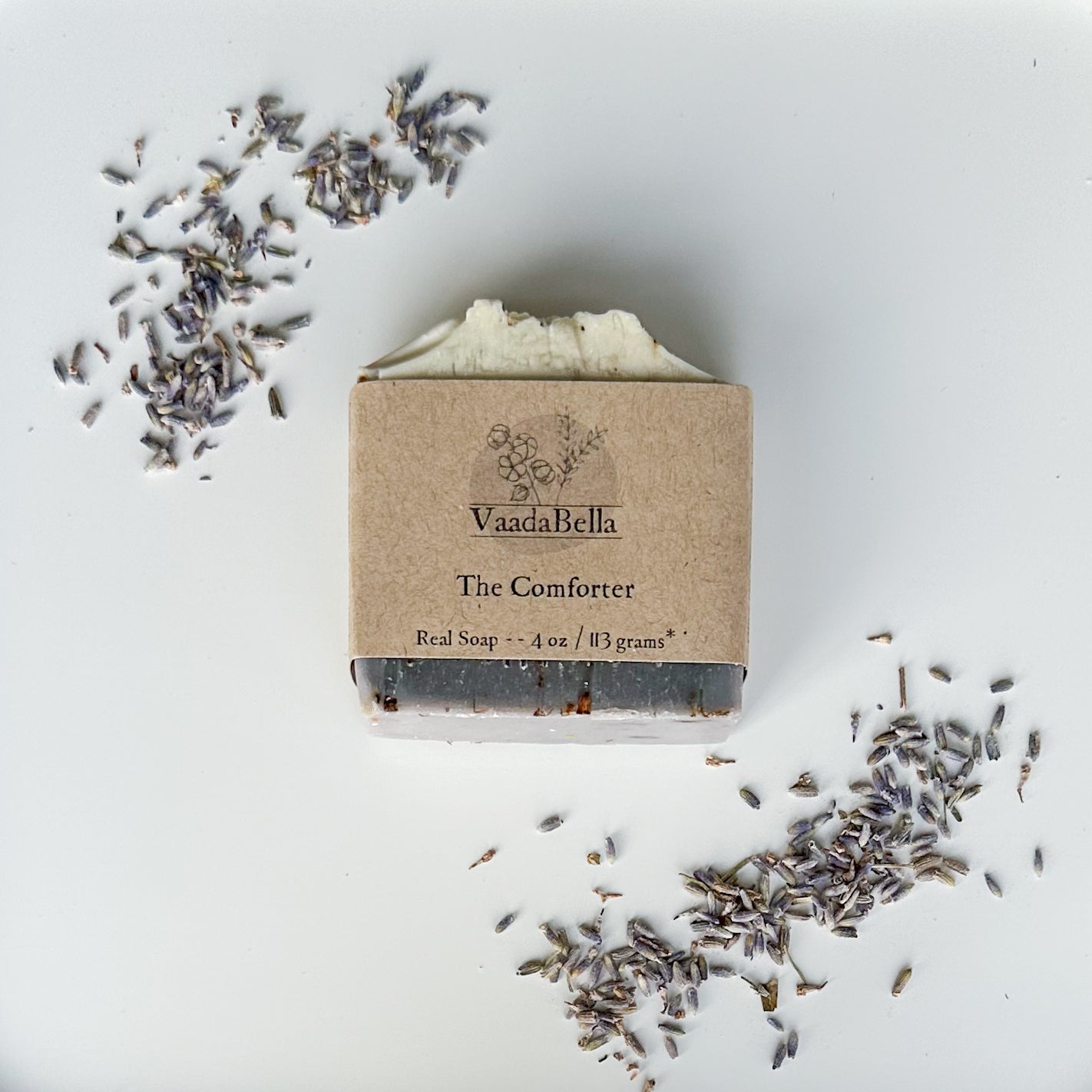 purple bottom, white top bar with kraft paper product sleve centered on bar.  Black ink prints the brand logo, "The Comforter" and "Real Soap -- 4 oz / 113 grams*".  French lavender buds arranged at the top left and bottom right of the image.