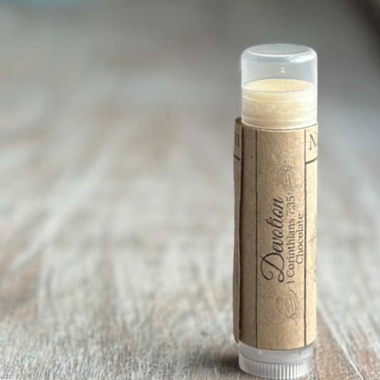 Stick of buttery yellow lip balm standing verticle on a weathered wood background, all on the right side of the image.  Kraft paper label with "Devotion", 1 Corinthians 7:35 and "chocolate" printed in black.