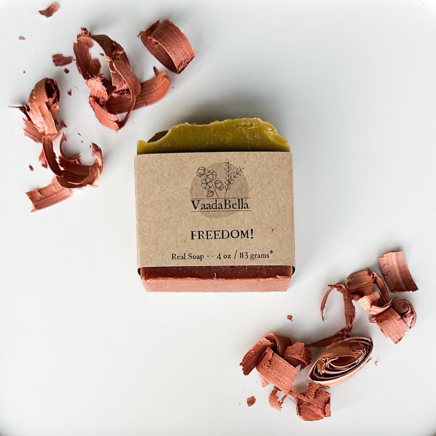 Green top, brick red bottom soap with brown kraft paper sleeve with black lettering and logo.  Cedar shavings at the top left and bottom right of the image.