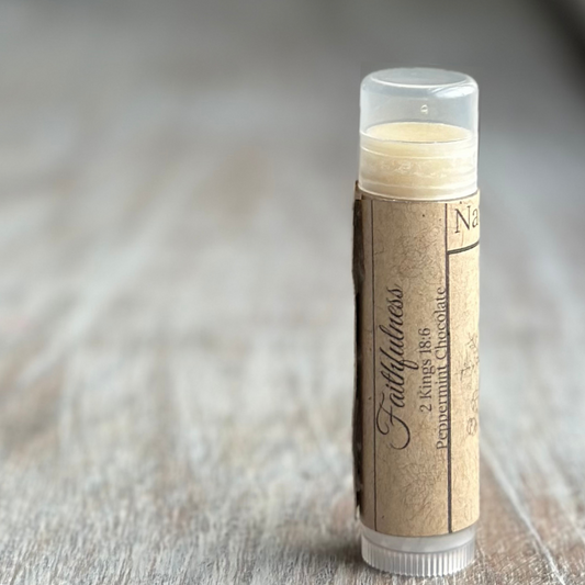 Stick of buttery yellow lip balm standing verticle on a weathered wood background, all on the right side of the image.  Kraft paper label with "Faithfulness", 2 Kings 18:6 and "peppermint chocolate" printed in black.