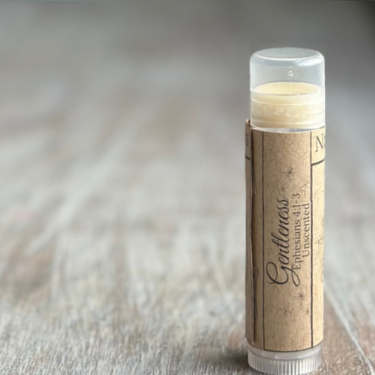 Stick of buttery yellow lip balm standing verticle on a weathered wood background, all on the right side of the image.  Kraft paper label with "Gentleness", Ephesians 4:1-3 and "unscented" printed in black.