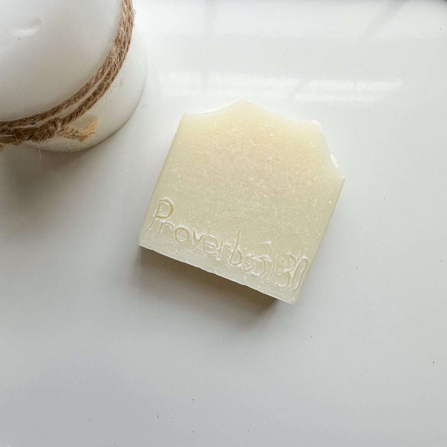 White bar with proverbs 31:30 etched on the front lays diagonal (upper right to lower left). Perspective: bird's-eye view.  White candle with hemp cord tied around the middle barely in the frame in the upper left
