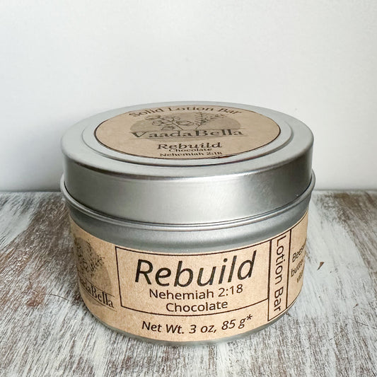 Front view of a circular silver metallic tin with lid.  brown kraft paper circle label centered. on the top, Logo, "Solid Lotion Bar" "Rebuild" "Nehemiah 2:18" "Chocolate" is all printed in black ink.  On the front, brown kraft paper sleeve wraps around the side of the tin with "Rebuild" "Nehemiah 2:18" "Chocolate" "Net wt. 3 oz, 85 g*" printed on it