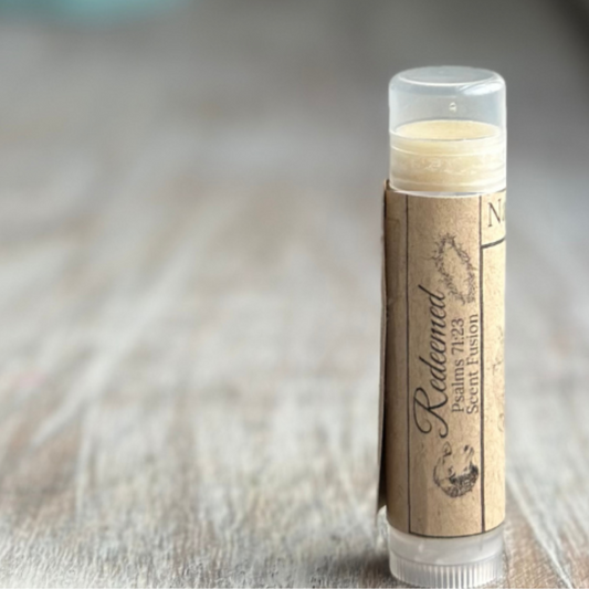 Stick of buttery yellow lip balm standing verticle on a weathered wood background, all on the right side of the image.  Kraft paper label with "Redeemed", Psalms 71:23 and "scent fusion" printed in black.