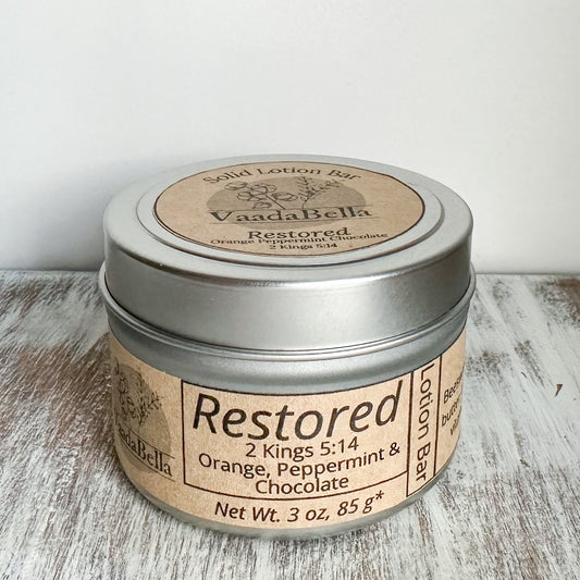 Front view of a circular silver metallic tin with lid.  brown kraft paper circle label centered. on the top, Logo, "Solid Lotion Bar" "restored" "Ruth 2:12" "Orange, Peppermint & Chocolate" is all printed in black ink.  On the front, brown kraft paper sleeve wraps around the side of the tin with "Restored" "Ruth 2:12" "Unscented" "Net wt. 3 oz, 85 g*" printed on it