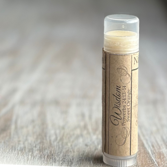 Stick of buttery yellow lip balm standing verticle on a weathered wood background, all on the right side of the image.  Kraft paper label with "Wisdom", Proverbs 24:13-14 and "chocolate" printed in black.